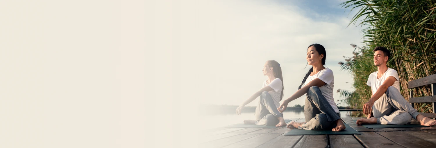 Three people practicing seated yoga poses on a wooden deck by a calm lake, embracing peace and mindfulness
